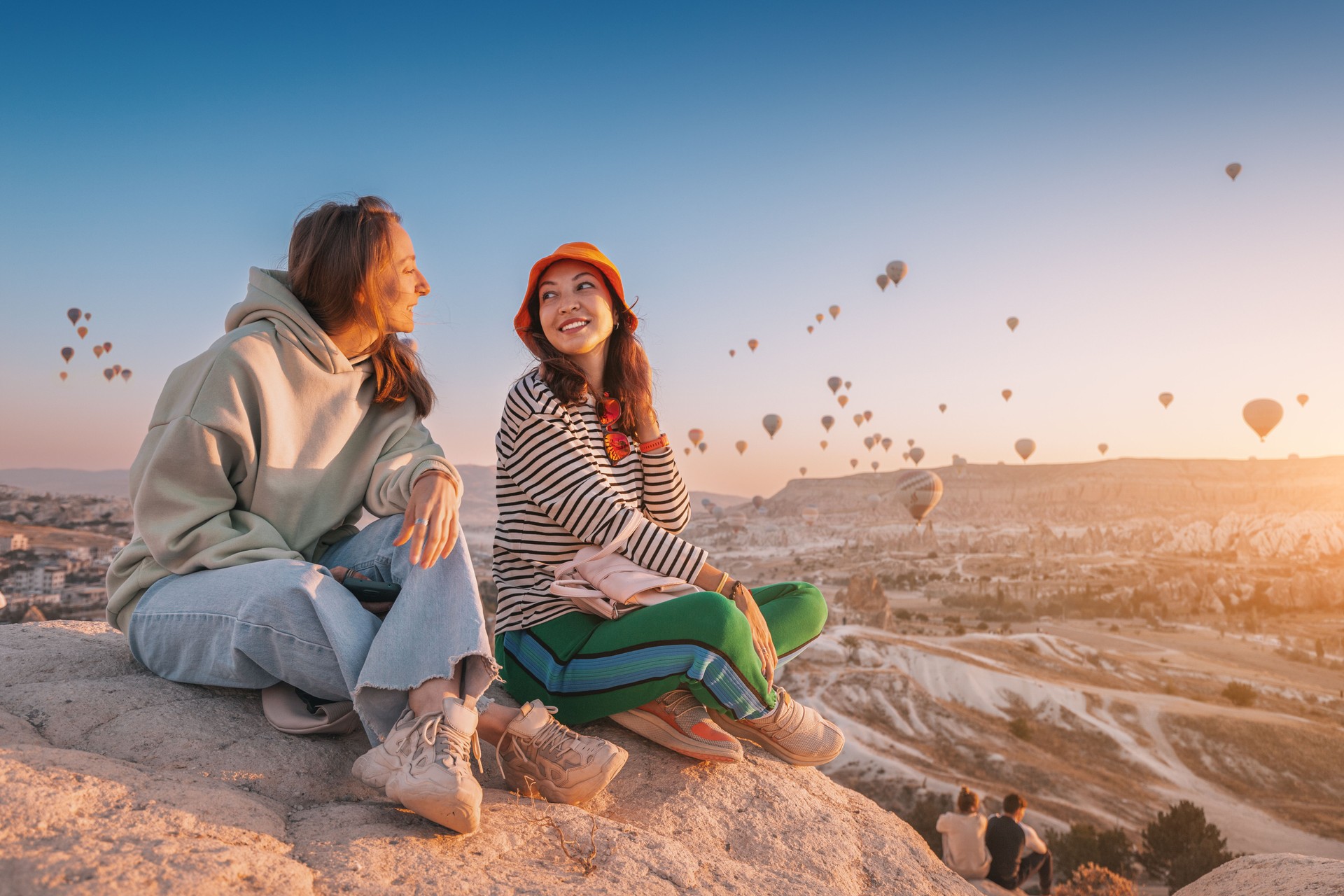 Freundinnen, die auf einem Aussichtspunkt auf einer Klippe sitzen und den Blick auf majestätische fliegende Heißluftballons in Kappadokien bewundern